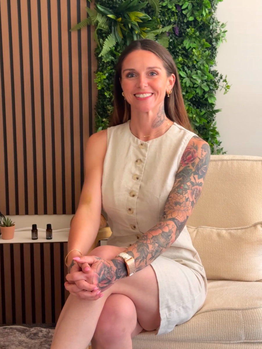 Portrait of Megan, founder of Rhythm Wellness, seated and smiling in her studio against a wall of greenery and warm wood paneling.